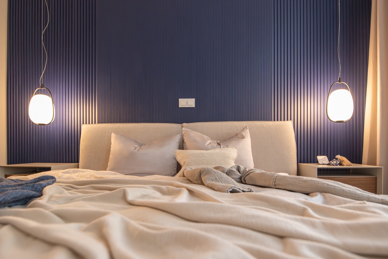 Schlafzimmer mit gepolstertem Kopfteil vor einer dunkelblauen Lamellenwand, links und rechts vom Bett je eine Pendelleuchte aus Glas über dem Nachtkästchen